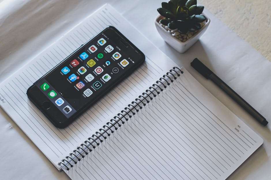 Smartfon z widocznymi ikonami aplikacji leży na otwartym notatniku obok czarnego długopisu.