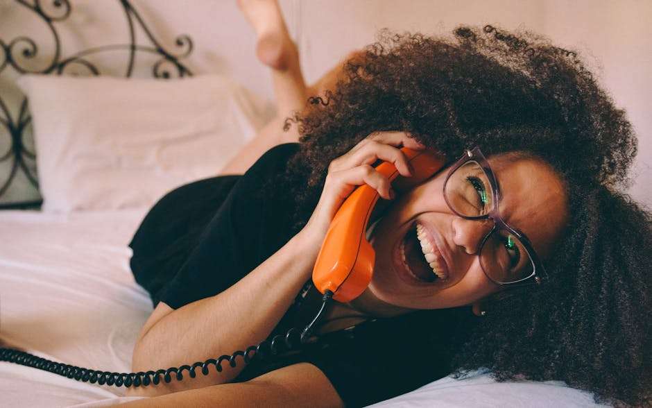 Kobieta z burzą kręconych włosów leży na łóżku, trzymając przy uchu pomarańczową słuchawkę telefonu.
