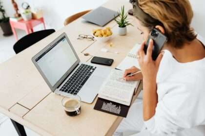 Kobieta przy biurku pracuje jednocześnie na laptopie, czyta książkę i rozmawia przez telefon.