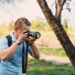 Mężczyzna w błękitnej koszulce fotografuje lustrzanką marki Sony w plenerze.
